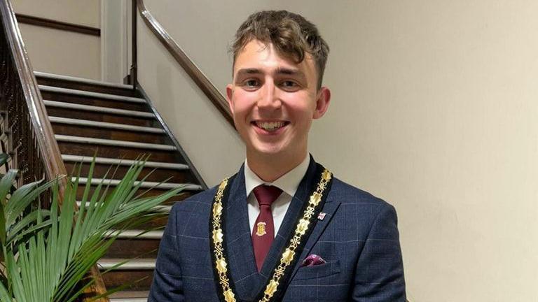 Harry Atkinson stood in front of a staircase, dressed in a suit, smiling and wearing his mayoral chain