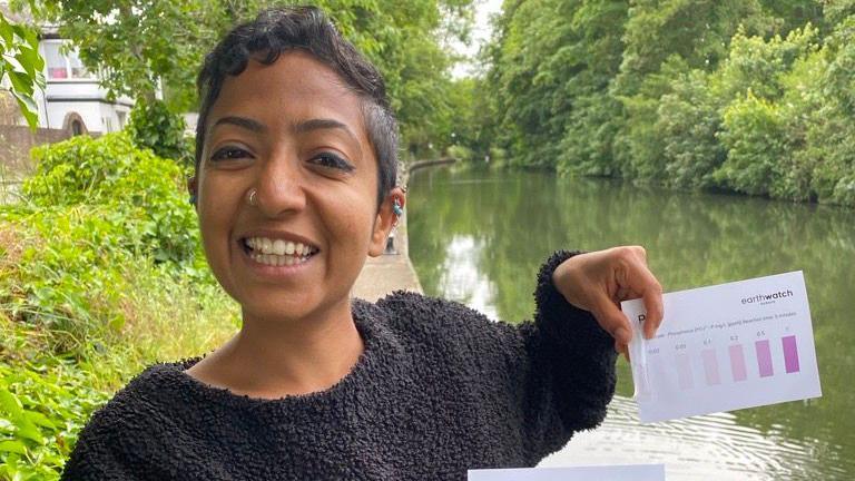 A woman standing on a canal towpath smiling and holding up two test cards for chemical levels in rivers. The canal is lined by trees with a house or pub on the left side.