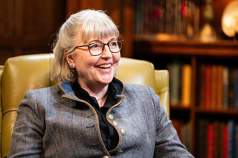 The Traitors' Lisa sitting in a yellow chair in the castle library , looking off to the left and smiling - she is wearing a grey herringbone jacket, black shirt and brown glasses. There are shelves full of books behind her.