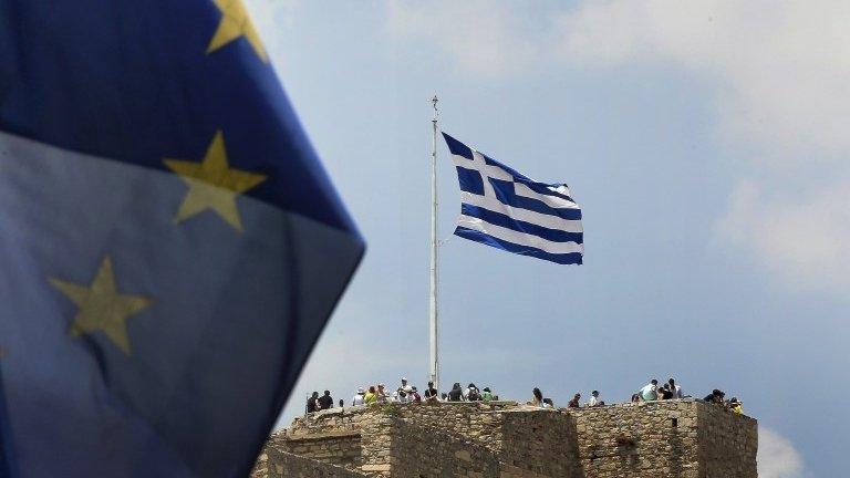 A European Union flag flutters as tourists gather around a Greek flag atop the Acropolis hill in Athen