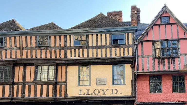 A row of tudor style buildings. They are orange, yellow and red with black panels on them. The yellow building in the middle has "Lloyd" written on it in capital letters
