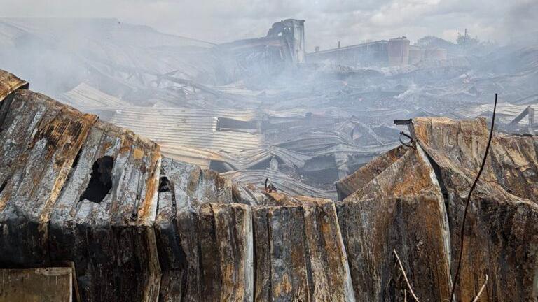 A burnt out building, showing burnt ground and burnt metal buildings, smoke in the air. 