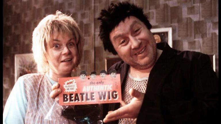 A still image from the Rab C Nesbitt TV show. Mary Doll stands next to Rab C, in his string vest and black jacket, and wearing a black wig.