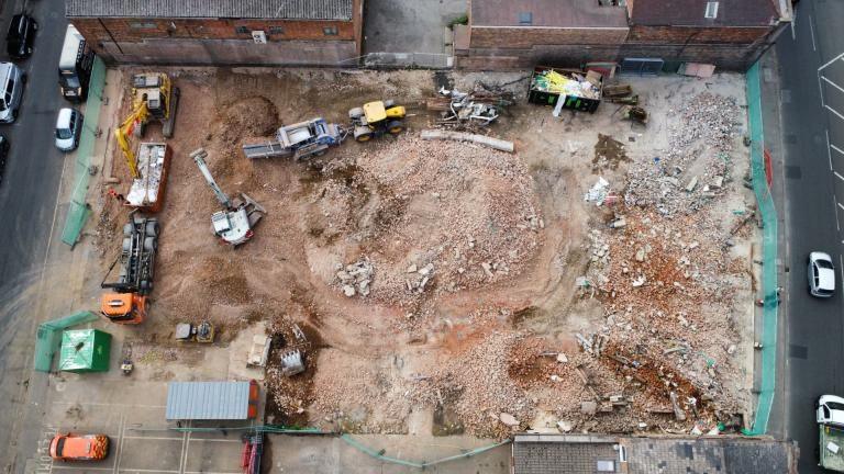 A bird's eye view of a construction area between two roads with six mechanical vehicles parked on it. There is also a skip and rubble on the site.