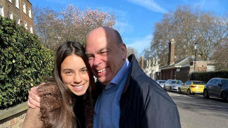 Hannah Lynch with long dark hair being hugged by Mike Lynch. They are both smiling at the camera and photographed in a street. 