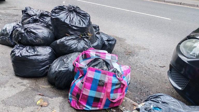 Roughly ten black bin bags piled up on a street, some on the pavement and some spilling onto the road.