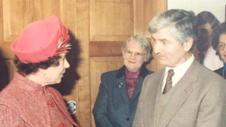 A young Queen Elizabeth on the left, wearing a pink jacket buttoned up and a matching pink hat with feathers. She is mid conversation with Stan Shaw, on the right, who has dark grey hair and is wearing a brown three-piece suit and white shirt.