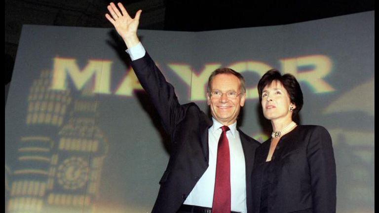 Jeffrey Archer and his wife Mary standing in front of a 'Mayor' backdrop. Jeffrey is wearing a dark suit and red tie and is waving. Mary is wearing a dark suit jacket and has short dark hair.