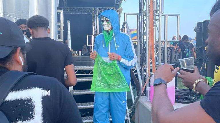 Takayo photographed next to a stage after his first performance at a music festival. He is wearing a blue tracksuit, a white mask and is holding a green scarf