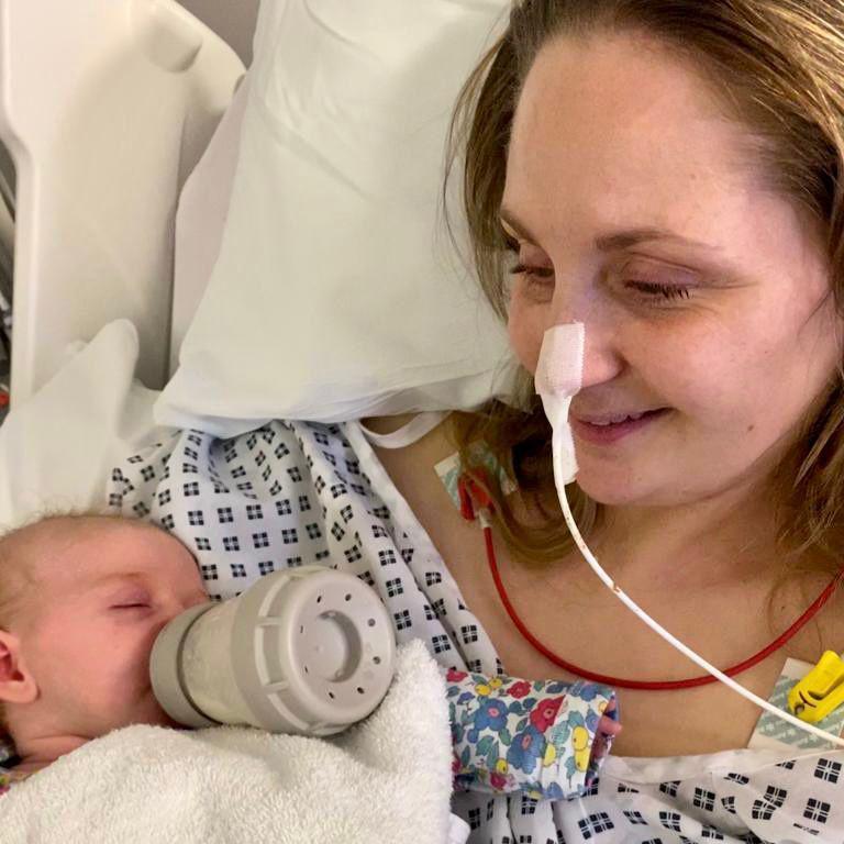 A woman in a hospital bed holding a baby, who is drinking from a bottle.