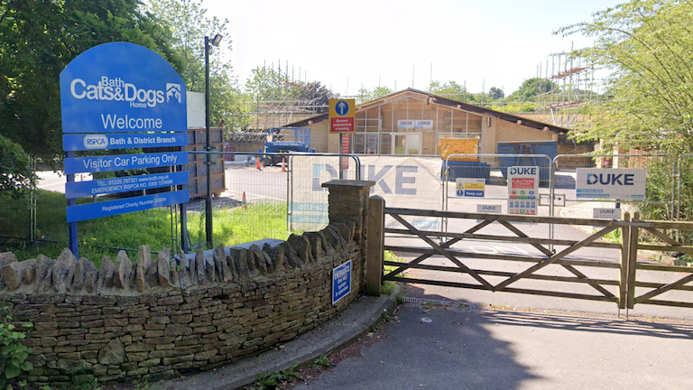 The exterior entrance to Bath Cats and Dogs Home in Somerset. There is a large blue sign above a curved brick wall which leads to a wide wooden gate. Behind it there is some metal fencing set up amid construction to convert an existing building into purpose built accommodation for animals.