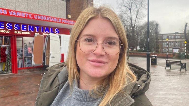 A woman with blonde hair, wearing glasses, is in the middle of a shopping street. There is a white van and a shop with red signage behind her on one side, and a bench and office building in the distance to the other side.