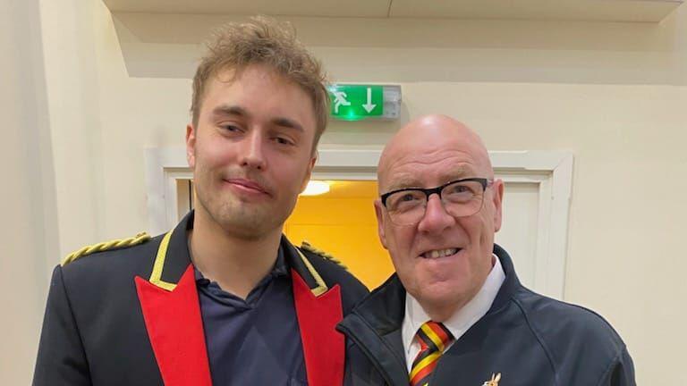 Sam Fender and Peter Lawson smile for the camera. Fender has short, light brown hair and is wearing a navy and red band uniform. Mr lawson is bald and is wearing glasses.