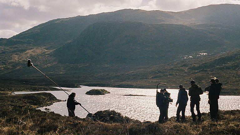 Filming taking place on the Isle of Harris - members of the crew are standing with cameras and equipment, near water that has the sun shimmering down on it. The hills and greenery can be seen in the distance and around them.