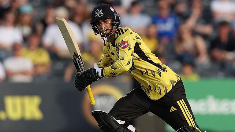 Kamran Dhariwal, wearing yellow and black and holding a bat, plays a shot while batting for Gloucestershire