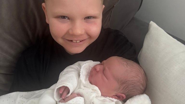 A little boy in a black shirt with a huge grin holds and baby in a white knitted sweater and swaddling sheets