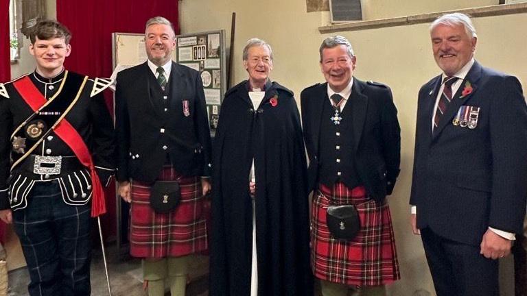 A group of pipers dressed in their kilts and tartan stand next to a vicar and another man in a suit with war medals in a church.