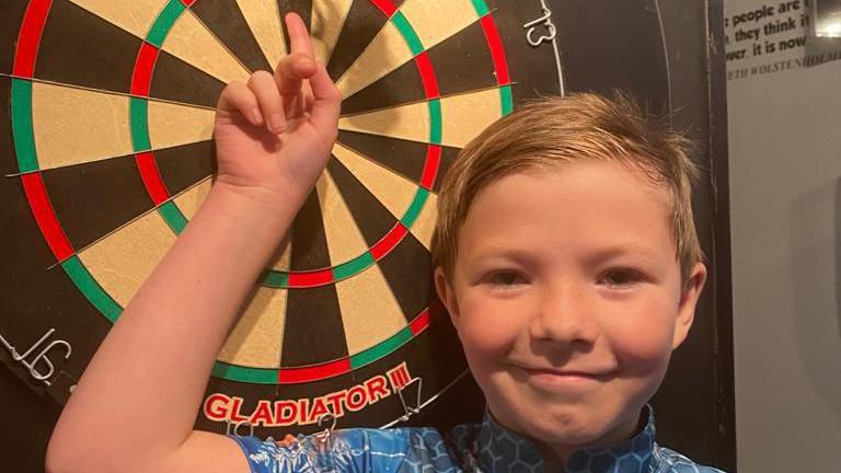 George wearing a blue top while smiling and pointing at a darts board.