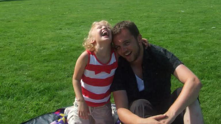 A man with short brown hair and stubble is sat on a blanket on grass with his knees up and hands around them and is wearing a dark top and shorts. He is laughing and leaning to the side where a young girl with curly blonde has her hand around him and head thrown back laughing. She is wearing a red and white striped top and beige trousers. 