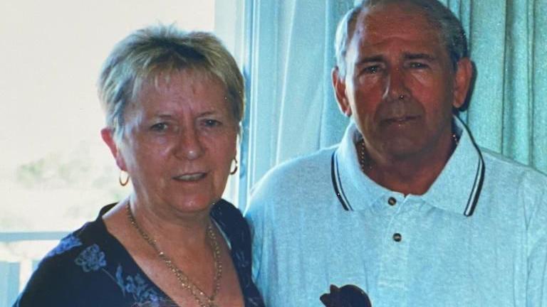 A woman with short blonde hair stands on the left. On her right is a man with short grey hair. Both are suntanned. The woman wears a dark blue top and a gold necklace. The man has a light grey polo shirt on.