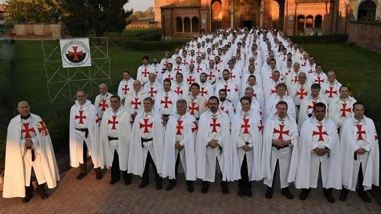 A large group of men stand in formation wearing white robes with red crosses on their chests. They are in front of a historic brick church, with a lawn and a banner displaying a similar cross emblem.