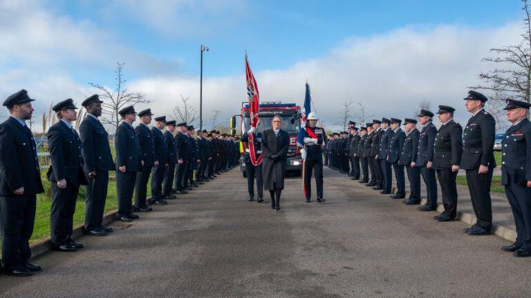 Colleagues dressed in formal suits and military-style hats line either side of a boulevard, while a fire engine drives slowly down the middle. In front of it, three people walk carrying two flags. There is a blue cloudy sky and grass either side of the road.