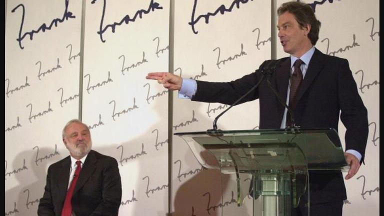 Tony Blair in a dark suit and brown tie stands at a podium with a 'Frank' themed background. Frank Dobson sits to his right wearing a dark suit and red tie.