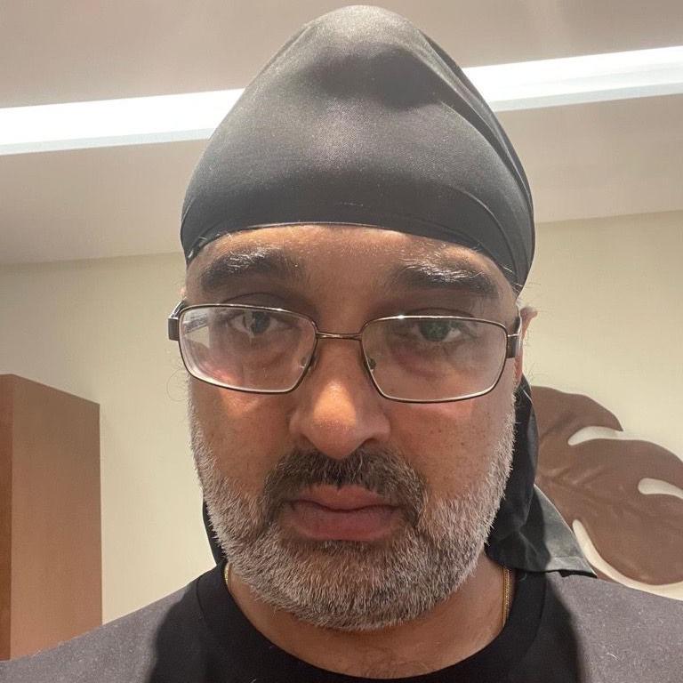 A selfie of a man in a black head scarf, and black T-shirt. He has black wire-framed glasses, a greying beard and a serious expression. Cream walls can be seen behind him.
