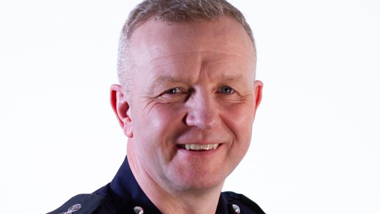 Neil Odin is wearing a blue uniform in front of a white background. He is smiling and has short, white hair 