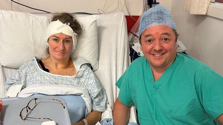 Susannah is propped up in a hospital bed with a bandage around her head. She is wearing a hospital gown and her glasses are lying on a table in front of her. Her surgeon, Iain Nixon, is kneeling beside her bed. He is wearing green scrubs and is smiling.