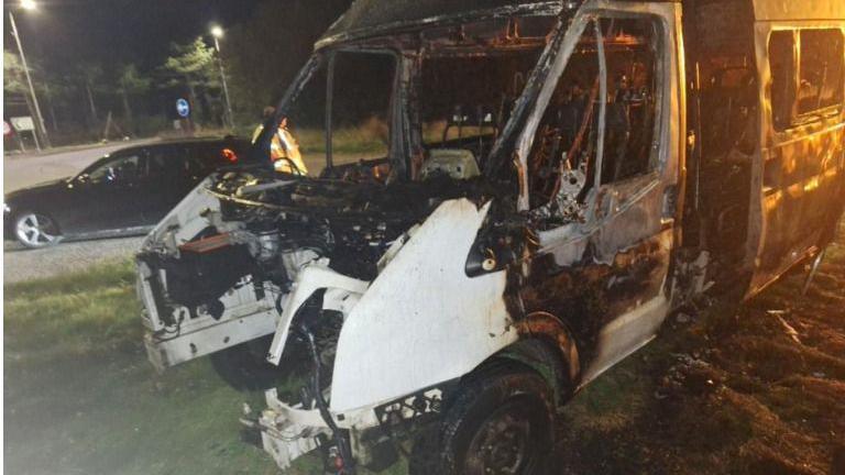 The front of the burnt-out minibus, showing how it has been completely destroyed. It is night time and there is one black car several yards behind it, and a person wearing high vis protective clothing. The windows, side door, bonnet and engine are missing.