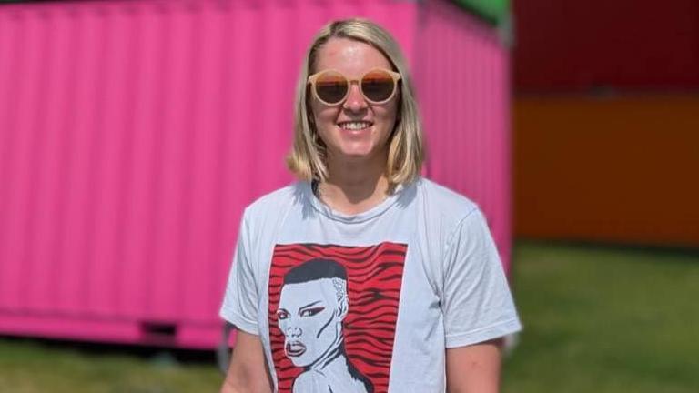 Cissy Badley. She has short blonde hair and is wearing brown sunglasses. She is smiling at the camera. A bright pink wall can be seen behind her.