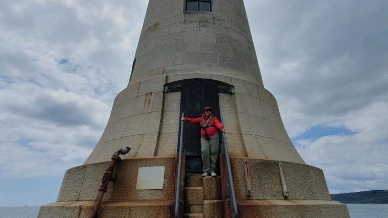 Jennifer Kloester to the right wearing a red coat, red cap and dark green trousers. She is wearing sunglasses and is stood on one of the alternating steps going up to the Plymouth breakwater lighthouse door. The lighthouse is light grey and has a window at the centre.