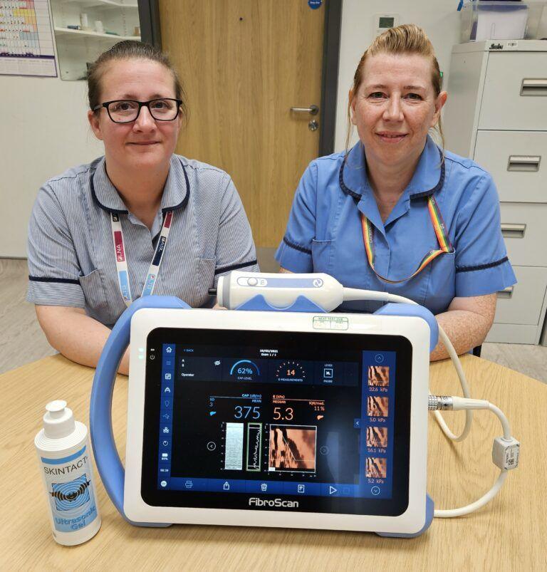 Two women sit at a table in medical uniforms with a fibroscan machine on the surface in front of them. The woman on the left of the image has dark hair in a pony tail, is wearing black framed glasses and is wearing a grey uniform. The woman on the right has light coloured hair and is wearing a blue uniform.