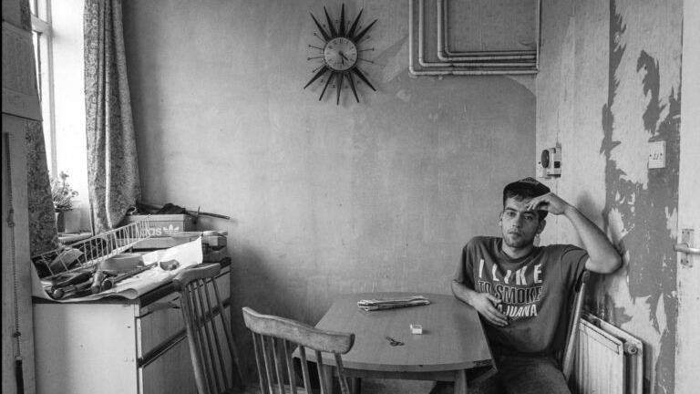 The black and white photo depicts a person seated at a small wooden table in a modest kitchen, surrounded by peeling paint, a sunburst wall clock, and a cluttered counter with dishes and cleaning supplies.