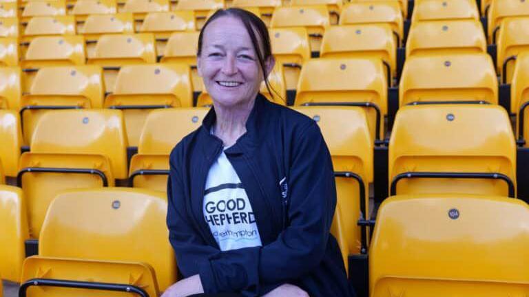 A woman with dark hair tied in a ponytail sits in a football stand that has gold-coloured seats. She is smiling and wearing a white T-shirt that reads "Good Shepherd", with a navy jacket with the same name on it.