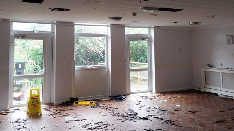 An abandoned building. There are holes in the ceiling panels and there is debris on the floor. There are yellow cleaning signs out on the floor.