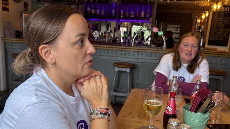 A woman with brown hair, tied back in a ponytail, sits with her chin on her folded hands. She is looking across a pub and has numerous charm bracelets on her right wrist. She is sat in a pub with another woman who has her arms crossed. 