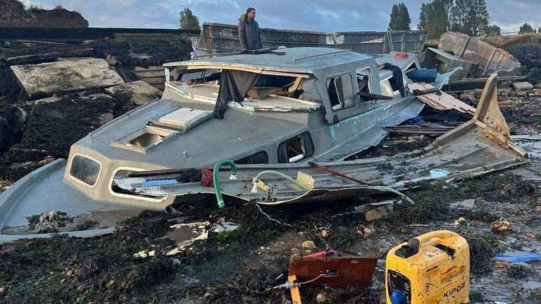 The wreckage of a houseboat. Most of the windows are smashed and fragments of the hull are lying on the ground. A man in a winter coat can be seen standing behind it.