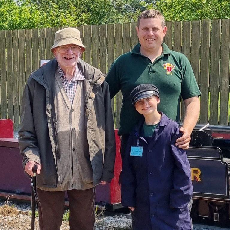 Two men and a boy stand outdoors in front of a wooden fence and a miniature train. On the left, an elderly man wearing a beige hat, glasses, checkered shirt, beige cardigan, and brown jacket holds a cane. In the middle, the man in a green polo shirt, who has an arm around his child - who is dressed in dark blue overalls and cap with an ID badge. Greenery and part of a miniature railway are visible in the background.