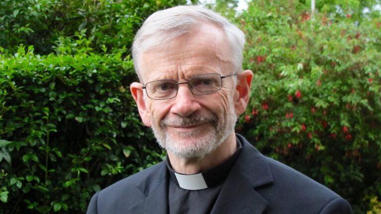 Man wearing glasses, a priest's collar and black jacket. He is standing outside with some hedges in the background, looking at the camera with a slight smile.