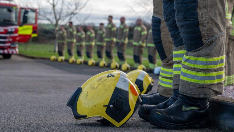 Colleagues line up to honour Gary Saville at the funeral, outside the crematorium. In the foreground we can see close ups of two yellow fire helmets on the ground, in front of the legs of two firefighters. They wear black boots and thick trousers with high viz strips at the hem. In the background is a fire engine and a further line of firefighters in their work outfits.