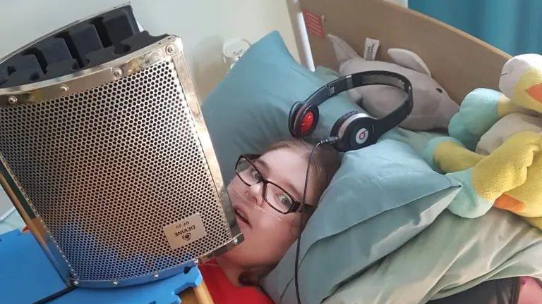 A woman lying in a bed with a large microphone placed in front of her and a set of headphones on her pillow