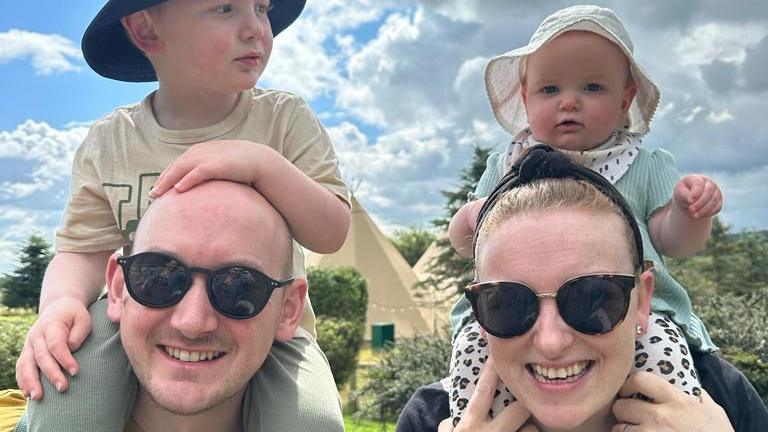 A man and woman, who wear sunglasses, carry two infants on their shoulders. They are outside on a sunny day.