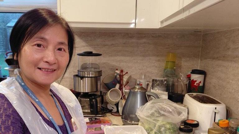 Poling Choi stands in a kitchen where she is working. She has a white plastic apron and a lanyard. The kitchen counter has a coffee machine, kettle, toaster, other utensils and food on it.