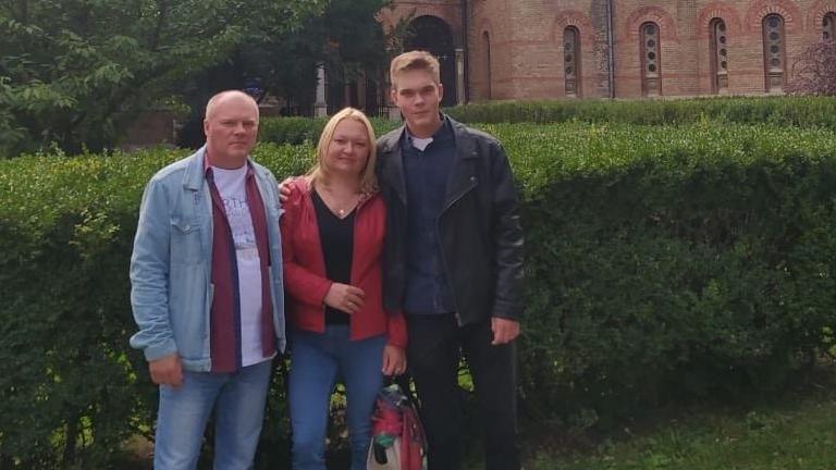 Illia with his mum and dad. He is wearing a black leather jacket, his mum has shoulder-length blonde hair and is wearing blue jeans and a red jacket and his dad has short fair hair and is wearing a denim short and jeans. They are standing in front of a green bush with an old building behind them. 