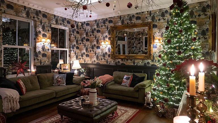 A living room with decorative wallpaper, a green sofa and a Christmas tree in the corner. There are candles and festive ornaments around the room. 