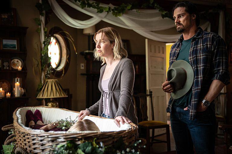 The image depicts a solemn scene involving a woman and a man in a dimly lit room with a wicker casket. A wicker casket is central in the image, adorned with flowers and containing what appears to be a body covered with a blanket. The woman, Lyle, is leaning on the casket, possibly in a moment of reflection or farewell. The man stands nearby, holding a hat and looking toward the first individual.