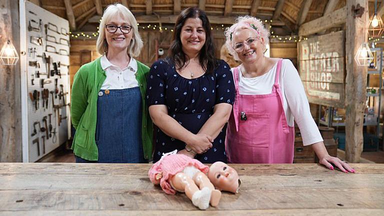 Julie Tatchell (left) in blue dungarees and green cardigan, Amanda Middleditch (right) with pink dungarees and white top, and Pip Martin in a blue dress stood behind a work bench with a damaged doll.