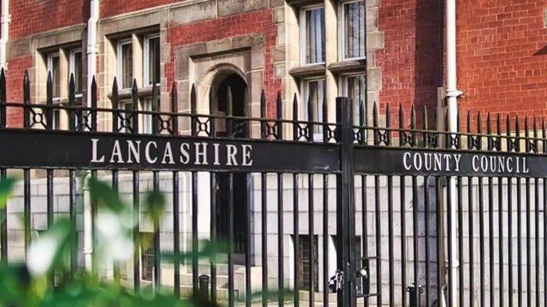 The black wrought iron gates at County Hall in Preston.  Lancashire County Council is written across them in white lettering.  The red-brick building can be seen behind them.  In the bottom left corner are some leaves from a tree.